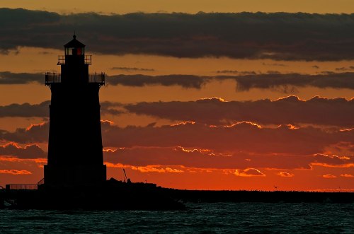 Silhouette of a lighthouse against a vibrant sunset with clouds reflecting warm colors over the water.