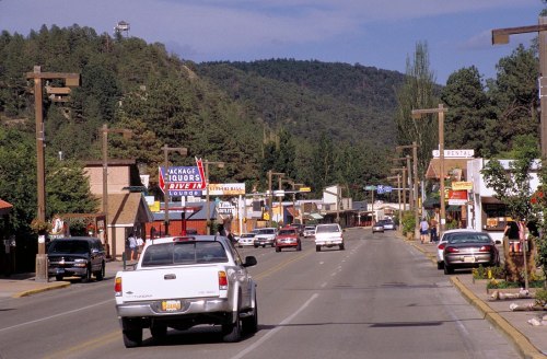 A small town street lined with shops and trees, with cars driving along and mountains in the background.