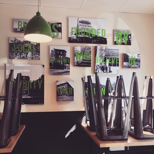 A cafe interior with stacked chairs and a wall of black-and-white photos featuring green text about local businesses.