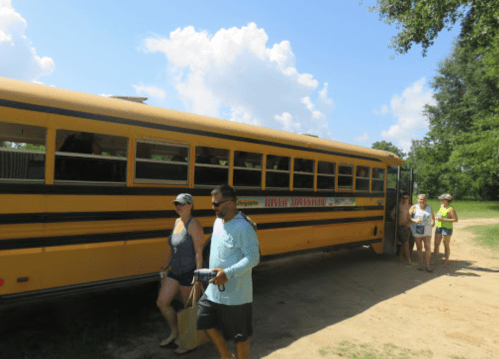 A yellow school bus parked on a dirt path with people getting on and off under a blue sky with fluffy clouds.