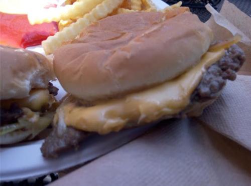 A close-up of a cheeseburger with melted cheese on a bun, served with fries in the background.