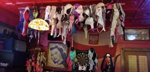 A colorful display of bras hanging from the ceiling in a bar, with vintage decor and a dartboard in the background.