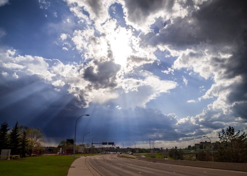 A dramatic sky with rays of sunlight breaking through dark clouds over a road lined with trees.