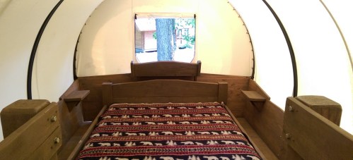 Cozy interior of a tent with a wooden bed and patterned blanket, surrounded by natural light.