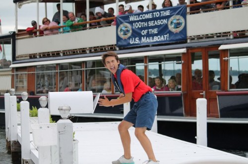 A young man in a red shirt poses playfully on a dock, with a crowd and a boat in the background celebrating the U.S. Mailboat's 100th season.
