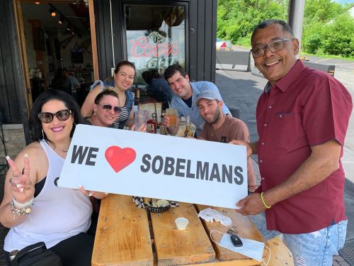 A group of six people at a table holding a sign that says "WE ❤️ SOBELMANS" with drinks and food around them.