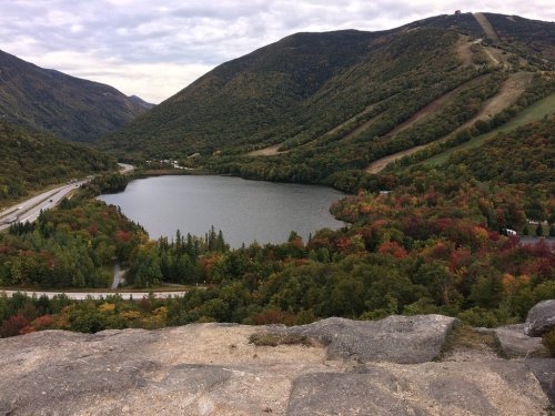 A scenic view of a lake surrounded by colorful autumn foliage and mountains under a cloudy sky.