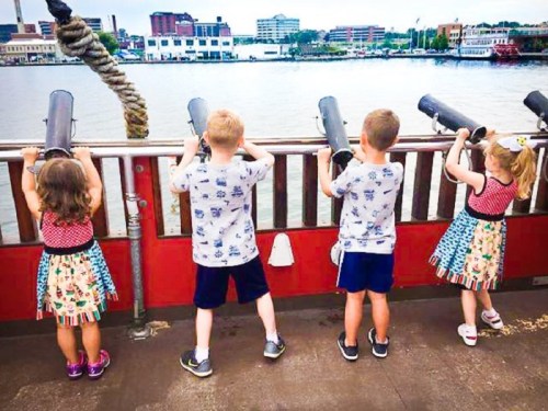 Four children on a boat, looking through binoculars at the water and city skyline in the background.