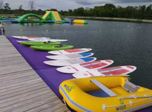 Colorful paddleboards and inflatable water toys lined up on a dock by a calm lake with greenery in the background.