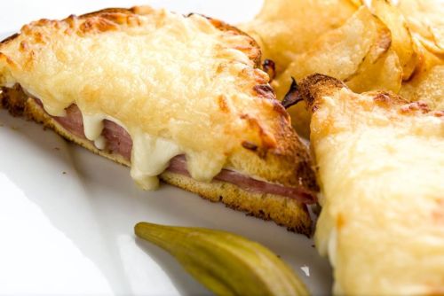 A close-up of a cheesy ham sandwich with crispy chips on the side, served on a white plate.