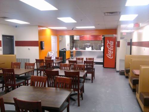 Interior of a casual dining restaurant with wooden tables, chairs, and a counter area featuring a Coca-Cola fridge.