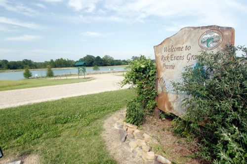 Sign welcoming visitors to Rick Evans Grand Prairie, with a scenic view of a lake and greenery in the background.
