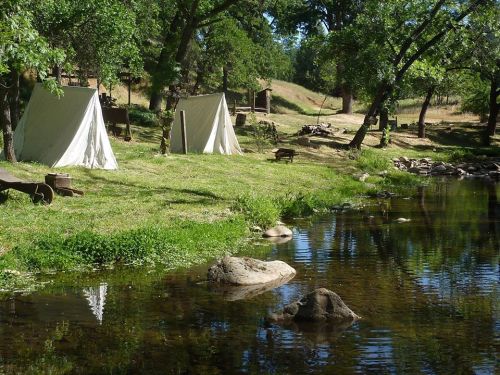 Two white tents by a calm river, surrounded by green grass and trees, with rocks visible in the water.