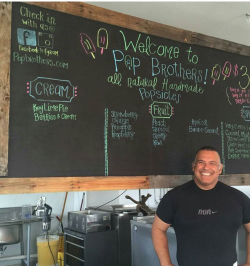 A smiling man stands in front of a chalkboard menu for Pop Brothers, featuring handmade popsicles and flavors.