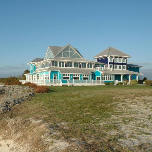 A large, colorful beach house with multiple levels and a gray roof, surrounded by grass and near the shoreline.