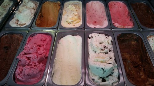A variety of colorful gelato flavors displayed in metal containers, including pink, white, brown, and beige options.