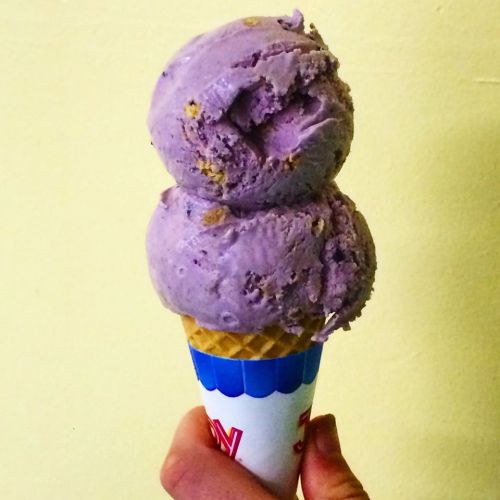 A hand holding a cone with two scoops of purple ice cream against a light yellow background.