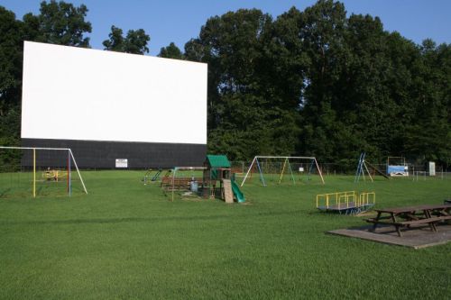 A grassy area featuring a large blank movie screen and a playground with swings and slides under a clear blue sky.