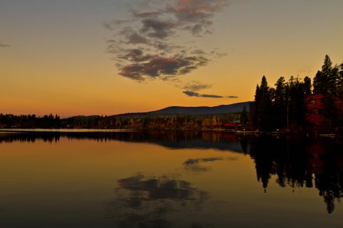 Sunset over a calm lake, reflecting trees and mountains, with a warm orange and purple sky.