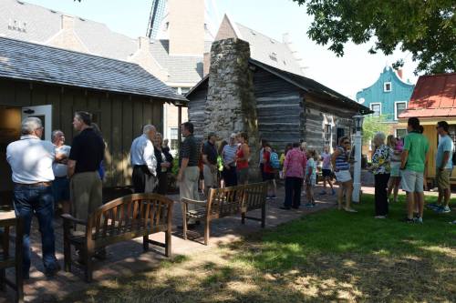 A group of people gathers outside historic buildings, chatting and enjoying a sunny day in a park-like setting.