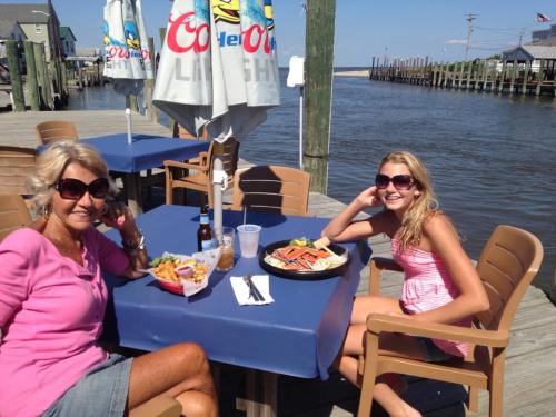 Two women sit at a table by the water, enjoying seafood and drinks on a sunny day.