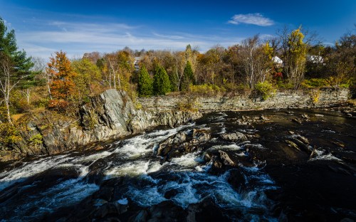 A scenic river flows over rocks, surrounded by autumn foliage and trees under a clear blue sky.