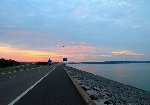 A lone runner jogs along a path beside a calm body of water at sunset, with colorful clouds in the sky.