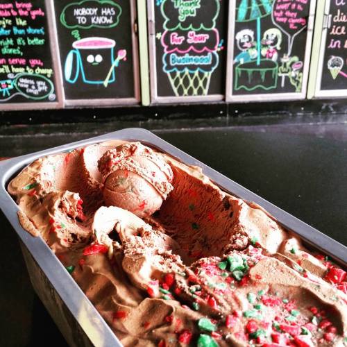 A tub of chocolate ice cream with red and green sprinkles, set against a colorful chalkboard backdrop.