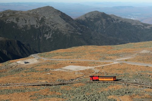 A red train travels along a winding track through a mountainous landscape with rocky terrain and distant hills.