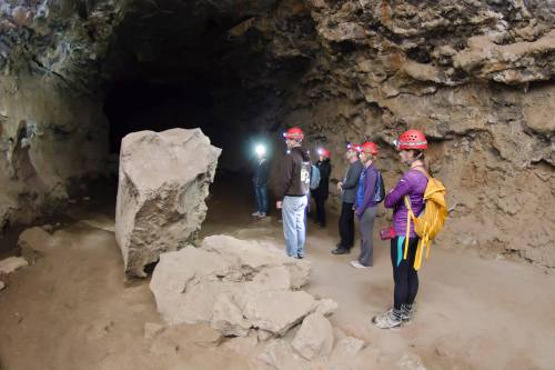 A group of people wearing helmets stands inside a cave, illuminated by headlamps, with rocky walls surrounding them.