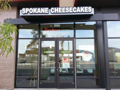 Exterior of Spokane Cheesecakes shop, featuring a large sign and glass doors with an "Open" sign visible.