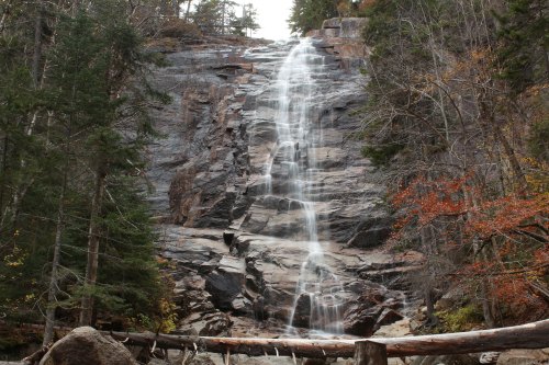 A cascading waterfall flows down a rocky cliff, surrounded by trees and autumn foliage.