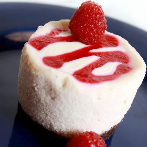 A creamy cheesecake topped with raspberry sauce and a fresh raspberry, served on a blue plate.