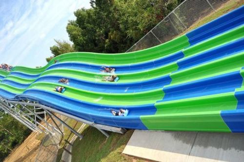 Aerial view of a colorful water slide with people sliding down on a sunny day, surrounded by trees.