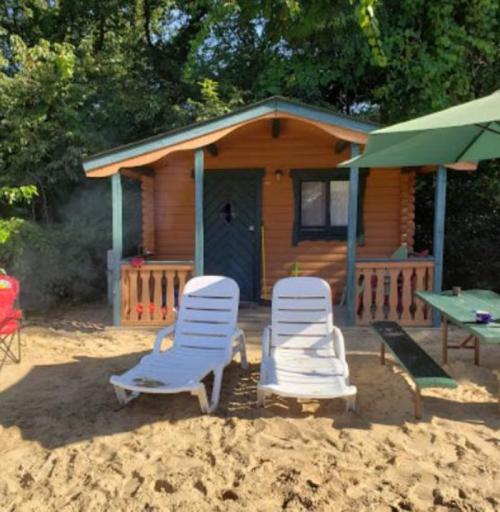 A cozy wooden cabin surrounded by trees, with two white lounge chairs and a green umbrella on sandy ground.
