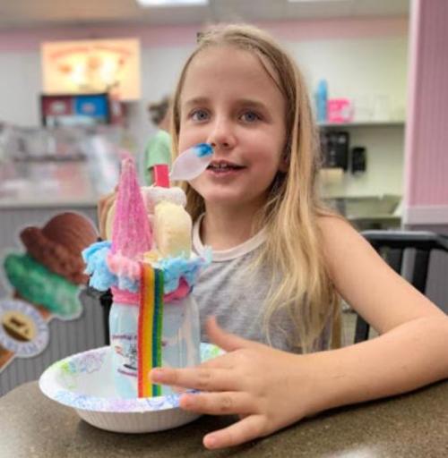 A young girl with long hair smiles while holding a colorful ice cream sundae in a café.