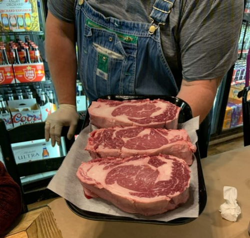 A person in overalls holds a tray with three large, marbled ribeye steaks.