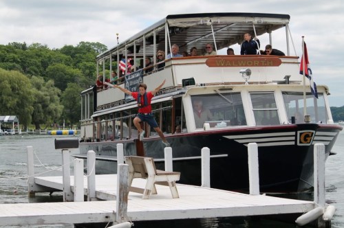 A boy in a red life jacket jumps off a dock towards a boat named "Walworth" with passengers on board.