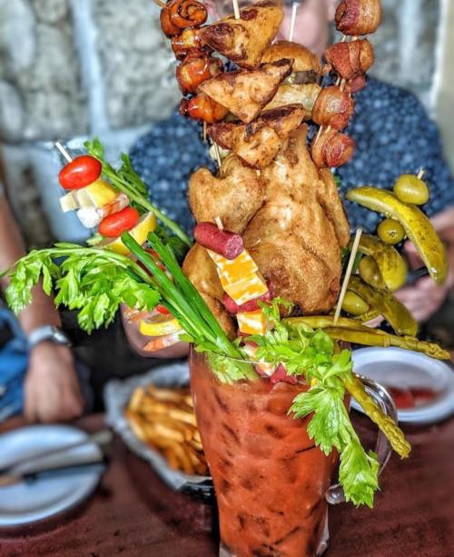 A towering Bloody Mary garnished with fried chicken, bacon, pickles, olives, and vegetables, served in a large glass.