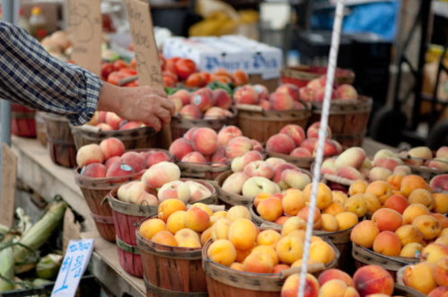 A hand reaches for fresh peaches among baskets of fruit at a market stall.