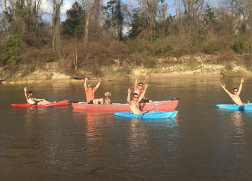 A group of five people in kayaks on a river, smiling and waving, surrounded by trees and a sandy bank.