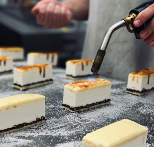 A chef uses a torch to caramelize the top of rectangular desserts on a tray, with a dusting of powdered sugar.