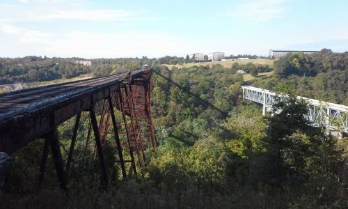 A rusted railway bridge spans a lush green valley, with a modern bridge visible in the distance under a clear blue sky.