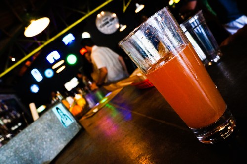 A glass of orange drink on a bar counter, with blurred patrons and colorful lights in the background.