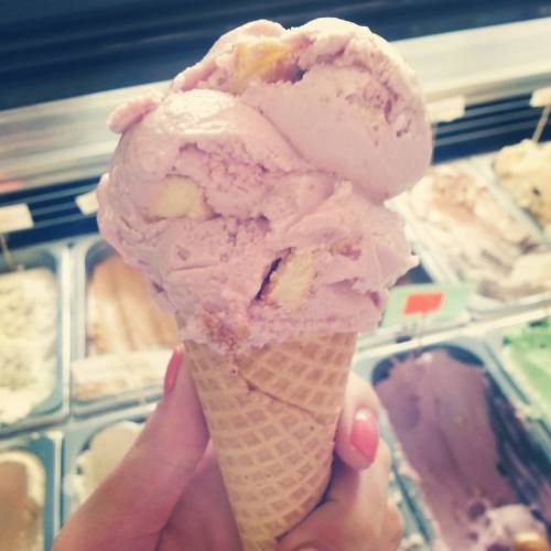 A hand holding a cone of pink ice cream with chunks, set against a background of various ice cream flavors.