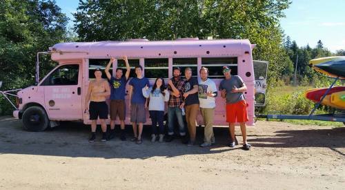 A group of eight people poses joyfully in front of a pink bus, surrounded by trees and kayaks.