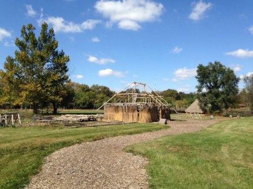 A rustic structure with a thatched roof surrounded by trees and a gravel path under a blue sky with clouds.
