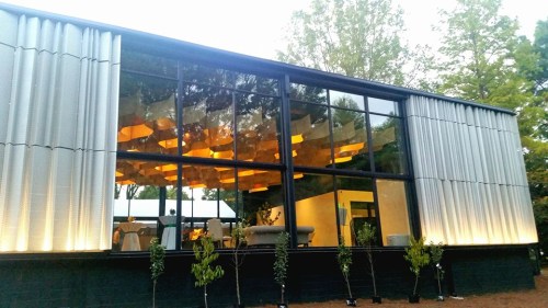 Modern building with large glass windows, featuring a stylish interior and illuminated ceiling, surrounded by greenery.