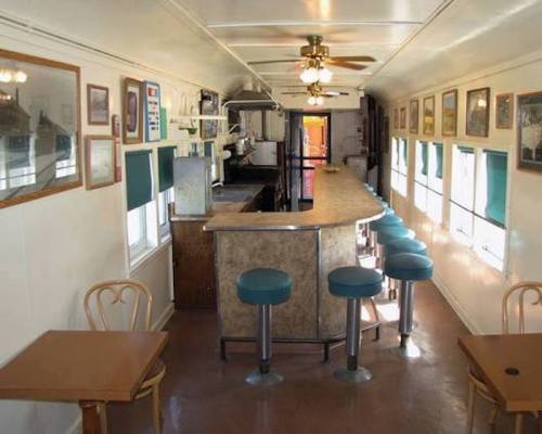 Interior of a vintage diner car featuring a curved bar, stools, and tables, adorned with framed pictures on the walls.
