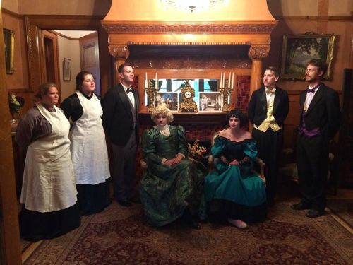 A group of people in vintage attire poses in a warmly lit room with a fireplace and decorative furnishings.